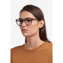 A woman with dark brown hair wearing tortoiseshell eyeglasses against a light gray background.