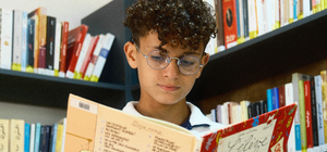 Young boy reading a book in a library with bookshelves in the background with optique et vision offer on it (Free lenses with frames purchased)