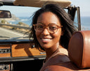 A woman wearing round eyeglasses and looking at the camera, with a pretty view behind her. 