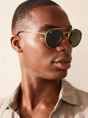 A close-up of a man wearing Ray-Ban aviator sunglasses with gold frames and dark green lenses, paired with a light beige linen shirt against a warm beige background.