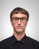 A man with short brown hair and glasses wearing a black button-down shirt, photographed against a light gray studio background.